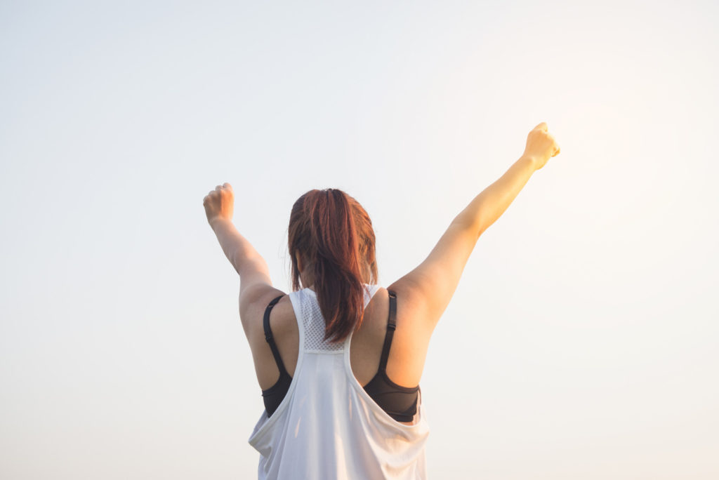Happy successful sportswoman raising arms to the sky on golden back lighting sunset summer. Fitness athlete with arms up celebrating goals after sport exercising and working out outdoors. Copy space.